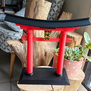 Könnte beinhalten: Ein rotes und schwarzes Torii-Tor-Modell mit schwarzem Sockel. Das Tor hat zwei rote Säulen und eine schwarze Oberseite mit einem goldenen Emblem in der Mitte. Das Modell wird auf einer Holzoberfläche ausgestellt, was auf einen Dekorationsartikel hindeutet.