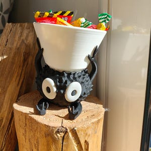 May include: A whimsical black creature-shaped candy bowl with large white eyes and a white bowl held above. The bowl is filled with colorful wrapped candies. The sculpture is placed on a wooden stump.