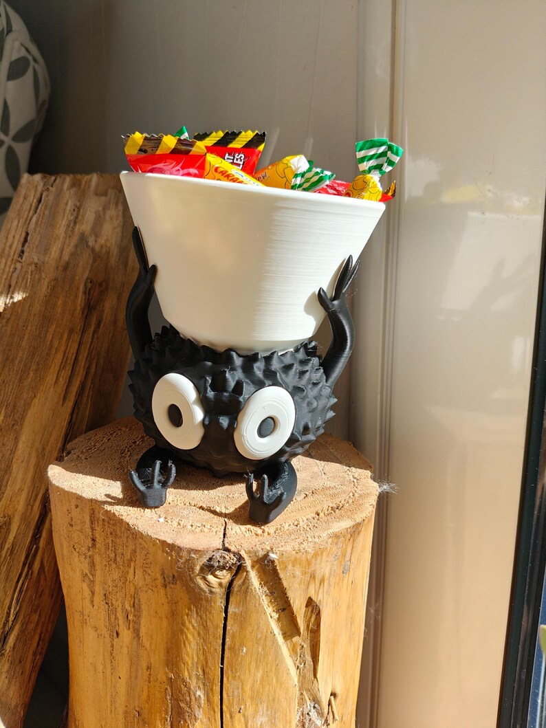 May include: A whimsical black creature-shaped candy bowl with large white eyes and a white bowl held above. The bowl is filled with colorful wrapped candies. The sculpture is placed on a wooden stump.