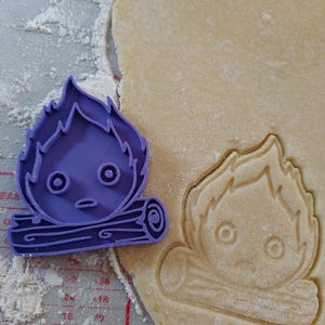 May include: A purple plastic cookie cutter in the shape of a cartoon campfire with a face, next to a cut-out of the same design in dough. The dough is on a gray surface with red measurement markings and flour.
