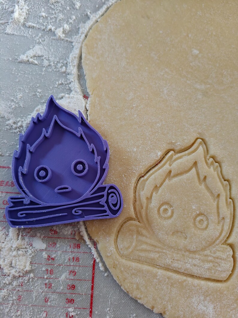 May include: A purple plastic cookie cutter in the shape of a cartoon campfire with a face, next to a cut-out of the same design in dough. The dough is on a gray surface with red measurement markings and flour.