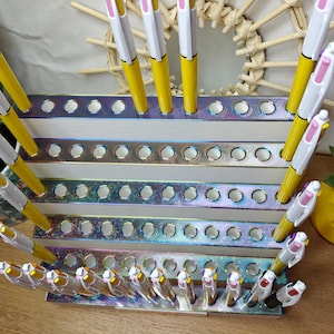 May include: A tiered, iridescent display stand with multiple circular cutouts holding numerous yellow and white multi-color ink pens. The stand features a metallic, rainbow-like finish on the front edges of each of the five horizontal levels.