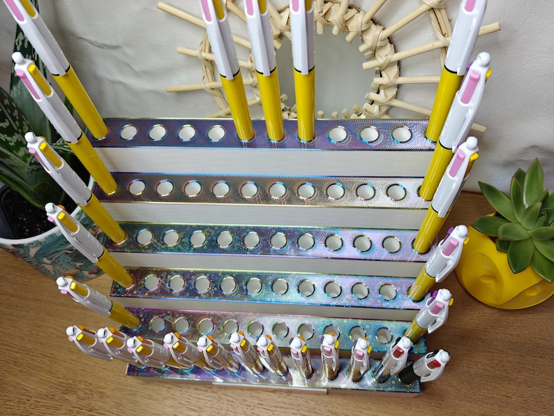 May include: A tiered, iridescent display stand with multiple circular cutouts holding numerous yellow and white multi-color ink pens. The stand features a metallic, rainbow-like finish on the front edges of each of the five horizontal levels.