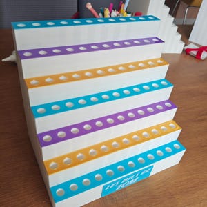 May include: A white tiered display stand with six levels, each featuring a row of circular holes for holding pens. The edges of the tiers alternate between teal, purple, and yellow. A teal rectangular label on the front reads LES BICS DE TOM.