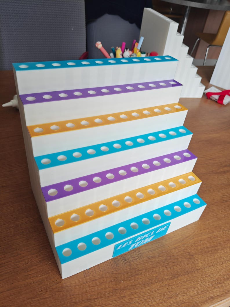 May include: A white tiered display stand with six levels, each featuring a row of circular holes for holding pens. The edges of the tiers alternate between teal, purple, and yellow. A teal rectangular label on the front reads LES BICS DE TOM.