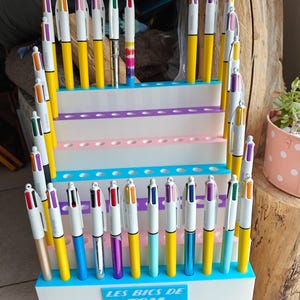May include: A tiered white display stand with blue accents holds numerous multi-color ballpoint pens. The base of the stand features a blue rectangular plaque with the white text "LES BICS DE TOM" printed in a bold, sans-serif font.