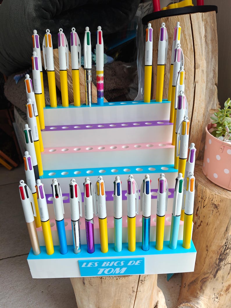 May include: A tiered white display stand with blue accents holds numerous multi-color ballpoint pens. The base of the stand features a blue rectangular plaque with the white text "LES BICS DE TOM" printed in a bold, sans-serif font.