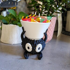 May include: A black, spiky, cartoon-like character holding a white bowl filled with wrapped candies. The character has large, oval white eyes and small arms and legs. The bowl is filled with colorful candy wrappers.