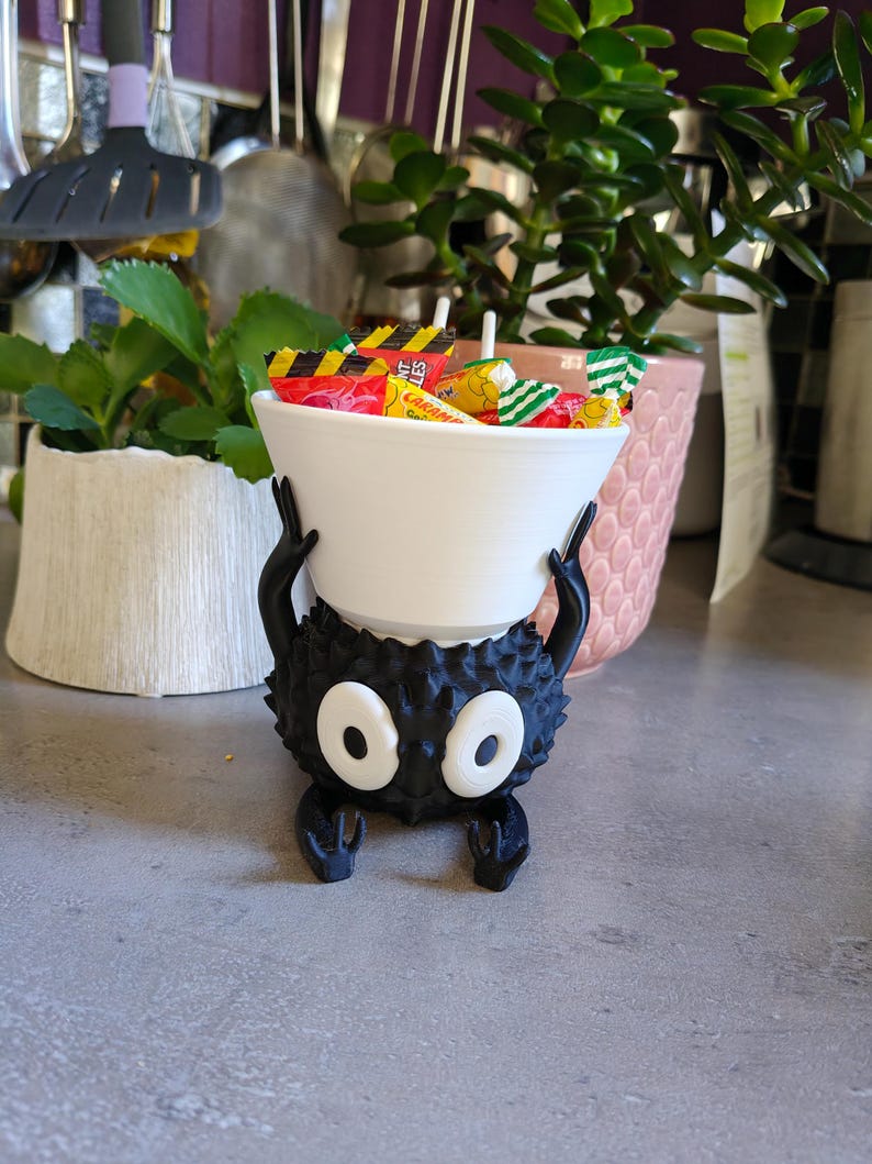 May include: A black, spiky, cartoon-like character holding a white bowl filled with wrapped candies. The character has large, oval white eyes and small arms and legs. The bowl is filled with colorful candy wrappers.