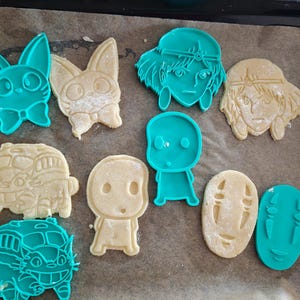 Peut inclure: Une collection d'emporte-pièces et de biscuits de différentes formes. Les emporte-pièces sont turquoise et les biscuits beiges. Les formes comprennent un lapin, un bus et plusieurs visages de personnages de dessins animés.