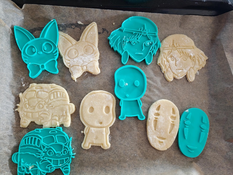 May include: A collection of cookie cutters and cookies in various shapes. The cutters are teal, and the cookies are beige. The shapes include a bunny, a bus, and several character faces, all from animated films.