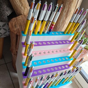 May include: A white tiered plastic display stand with multiple circular holes, holding numerous multi-colored retractable ballpoint pens. The stand features horizontal stripes in shades of blue, purple, and pink, designed to organize and showcase pens.