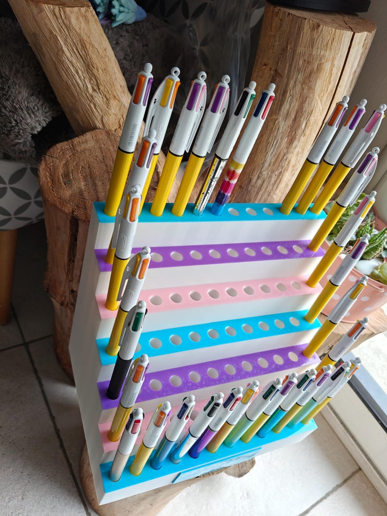 May include: A white tiered plastic display stand with multiple circular holes, holding numerous multi-colored retractable ballpoint pens. The stand features horizontal stripes in shades of blue, purple, and pink, designed to organize and showcase pens.