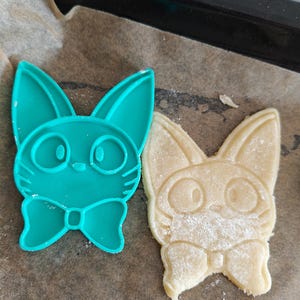 May include: A teal plastic cookie cutter in the shape of a cat with a bow tie next to a baked cookie in the same shape. The cookie is light brown and dusted with flour. The items are on parchment paper.
