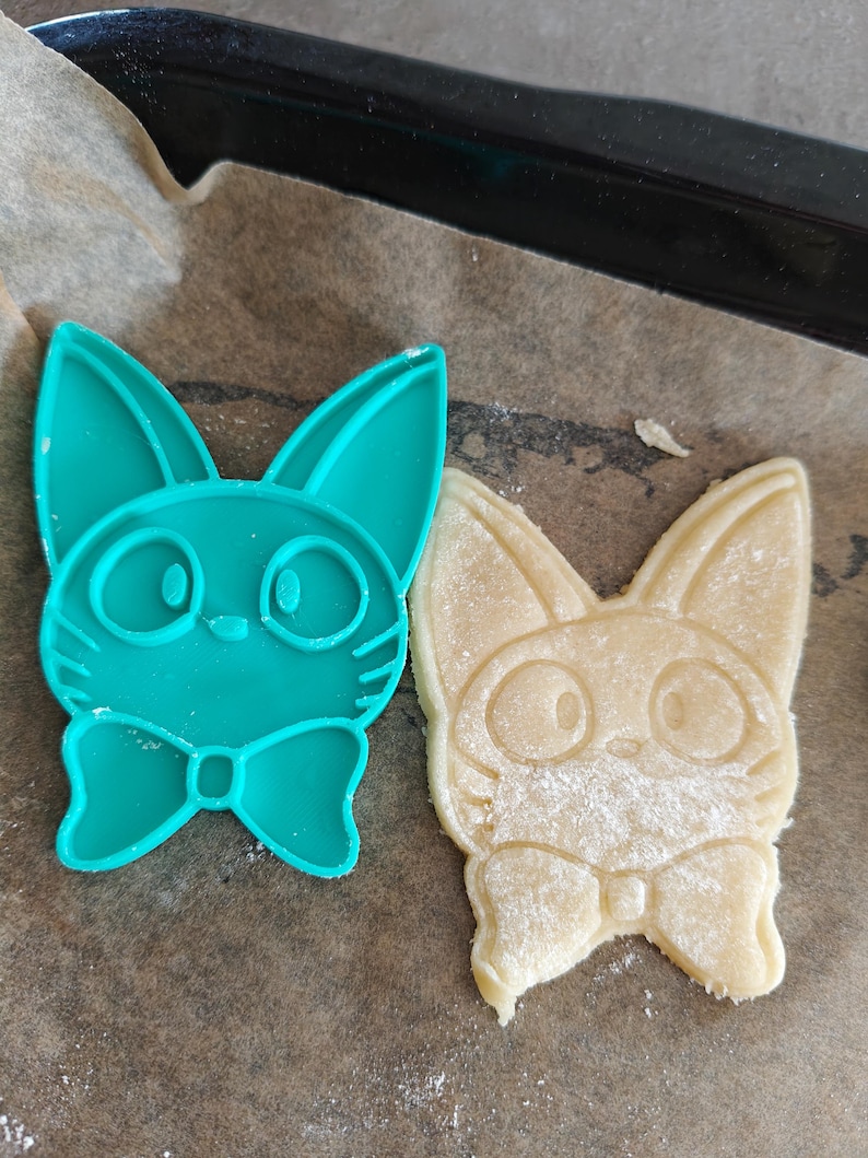 May include: A teal plastic cookie cutter in the shape of a cat with a bow tie next to a baked cookie in the same shape. The cookie is light brown and dusted with flour. The items are on parchment paper.