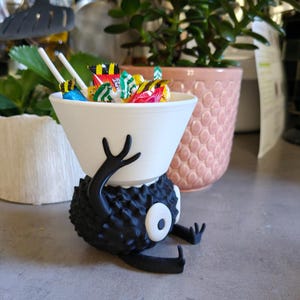 May include: A whimsical, 3D-printed candy bowl in the shape of a cartoon character. The white bowl is filled with colorful wrapped candies. The character is black with a spiky texture, white eye, and outstretched arms.