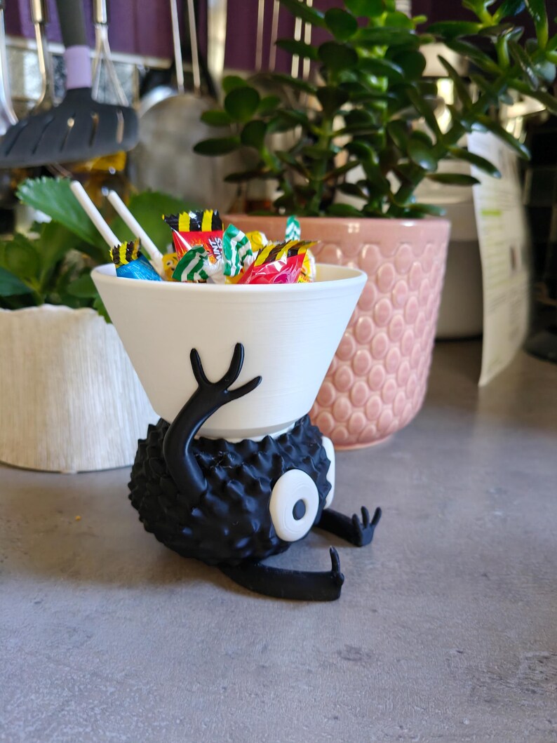 May include: A whimsical, 3D-printed candy bowl in the shape of a cartoon character. The white bowl is filled with colorful wrapped candies. The character is black with a spiky texture, white eye, and outstretched arms.