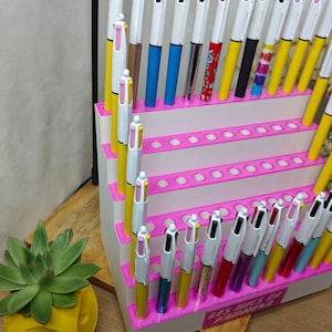 May include: A white tiered display stand with bright pink edges, holding numerous multi-color ink pens. The pens feature white barrels with various colored lower sections and clips. The base of the stand displays the text Les Bics de Fabrina.