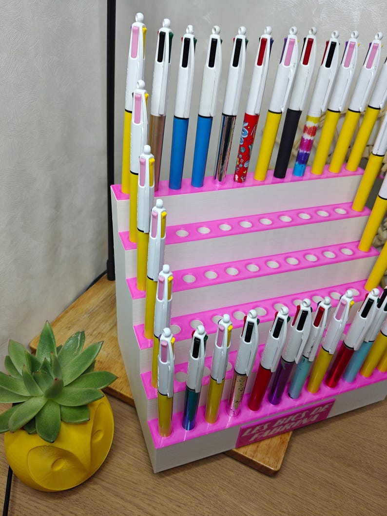 May include: A white tiered display stand with bright pink edges, holding numerous multi-color ink pens. The pens feature white barrels with various colored lower sections and clips. The base of the stand displays the text Les Bics de Fabrina.