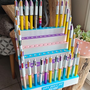 May include: A white tiered display stand holding numerous multi-colored ballpoint pens. The stand features rows of circular holes for pen storage and a blue rectangular sign at the base with the white text LES BICS DE TOM printed on it.