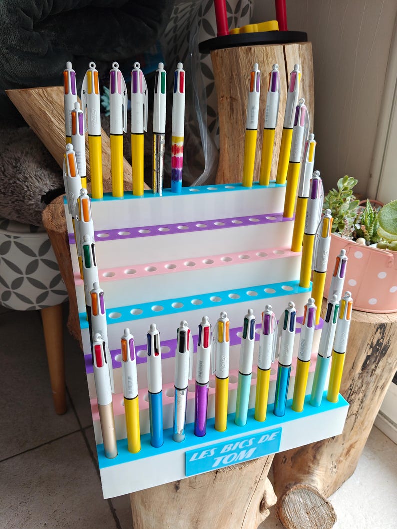 May include: A white tiered display stand holding numerous multi-colored ballpoint pens. The stand features rows of circular holes for pen storage and a blue rectangular sign at the base with the white text LES BICS DE TOM printed on it.
