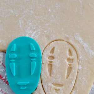 May include: A teal plastic cookie cutter in the shape of a character's face from the animated film, Spirited Away, next to a cutout of the same shape in dough. The dough is dusted with flour and sits on a red and white striped surface.