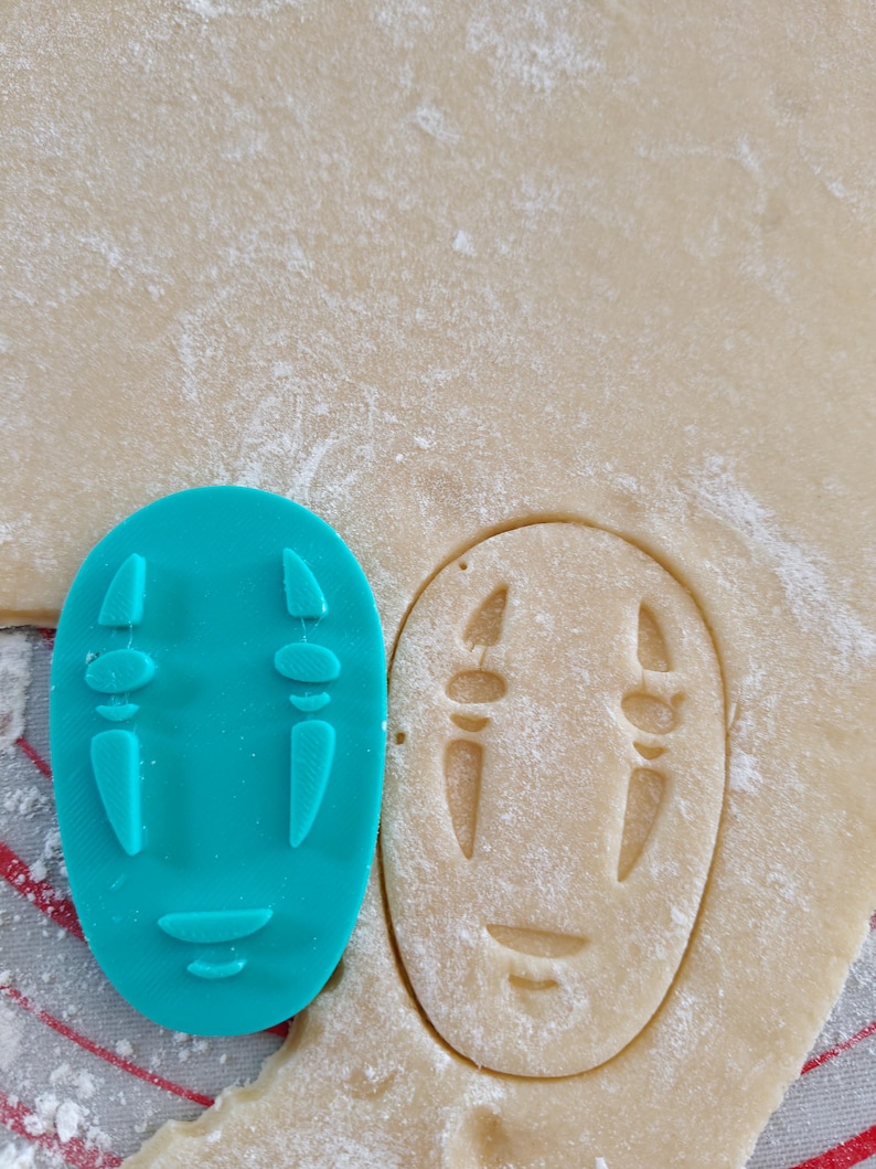 May include: A teal plastic cookie cutter in the shape of a character's face from the animated film, Spirited Away, next to a cutout of the same shape in dough. The dough is dusted with flour and sits on a red and white striped surface.