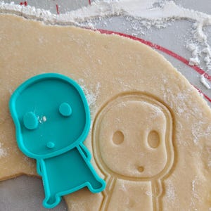 May include: A teal plastic cookie cutter in the shape of a cartoon character sits next to a cutout of the same character in dough. The dough is on a red and white silicone baking mat, with flour sprinkled around.
