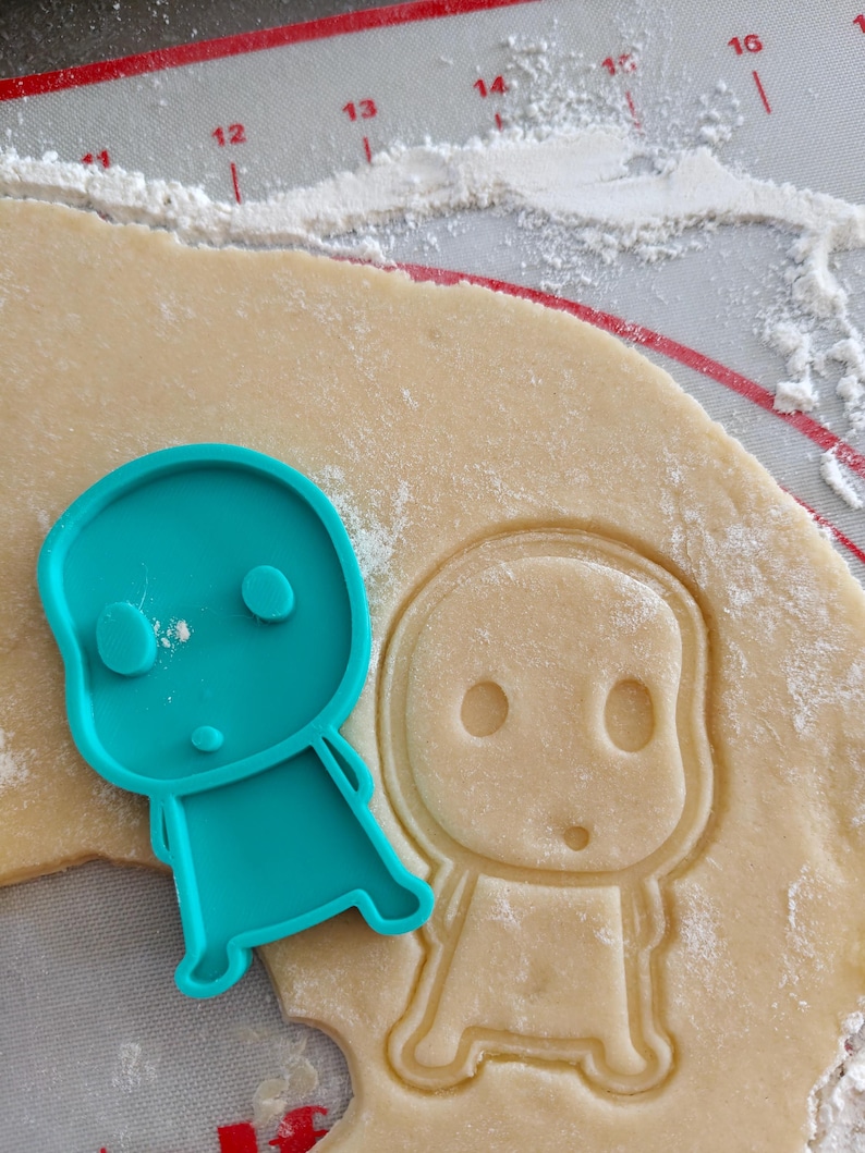 May include: A teal plastic cookie cutter in the shape of a cartoon character sits next to a cutout of the same character in dough. The dough is on a red and white silicone baking mat, with flour sprinkled around.