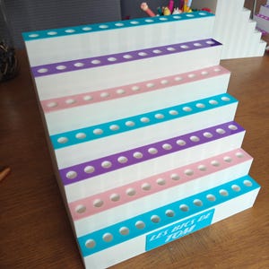 May include: A white tiered display stand with six levels, each featuring a row of circular holes for holding items. The edges of the tiers are accented with alternating teal, purple, and pink stripes. A teal label reads LES BICS DE TOM.