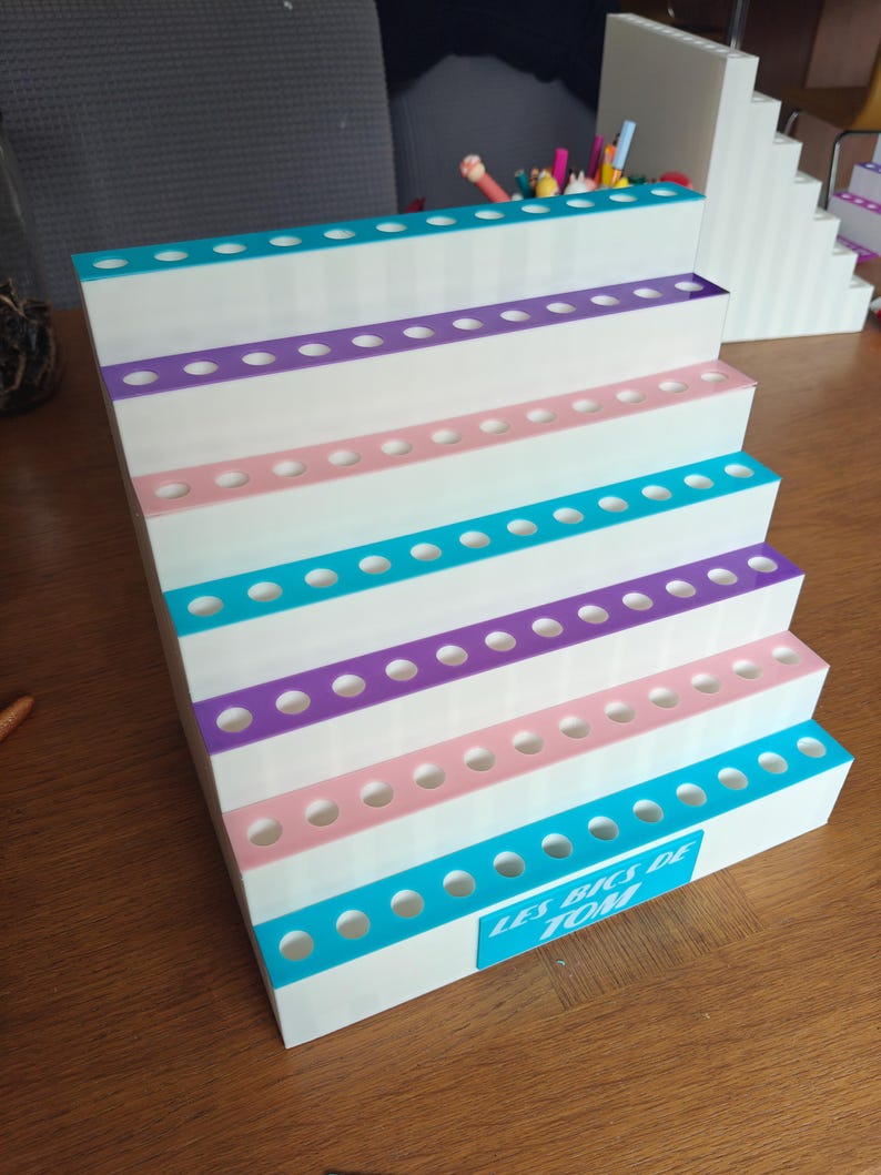 May include: A white tiered display stand with six levels, each featuring a row of circular holes for holding items. The edges of the tiers are accented with alternating teal, purple, and pink stripes. A teal label reads LES BICS DE TOM.