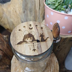 Peut inclure: Un bocal en verre transparent avec un couvercle en bois gravé d'une illustration d'une fille et d'un chat. Le couvercle a une poignée en bois. Le bocal est posé sur une base en bois, avec une jardinière rose à pois en arrière-plan.