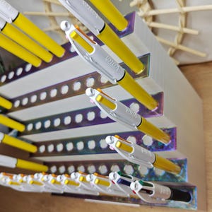 May include: A white tiered pen organizer with iridescent accents holding multiple yellow and white multi-color ballpoint pens. The organizer features rows of circular cutouts designed to hold the pens upright in a stepped, angled display configuration.
