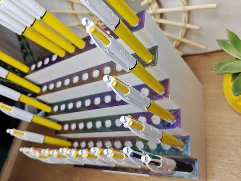 May include: A white tiered pen organizer with iridescent accents holding multiple yellow and white multi-color ballpoint pens. The organizer features rows of circular cutouts designed to hold the pens upright in a stepped, angled display configuration.