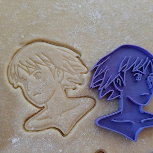 May include: A purple plastic cookie cutter in the shape of a person's profile, next to a cookie cut out of the same shape. The cookie is in a light brown dough.