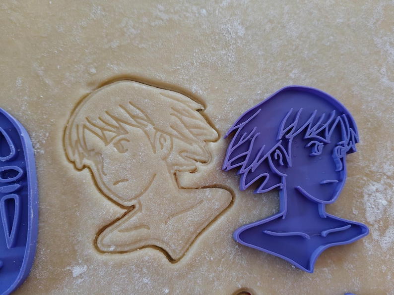 May include: A purple plastic cookie cutter in the shape of a person's profile, next to a cookie cut out of the same shape. The cookie is in a light brown dough.