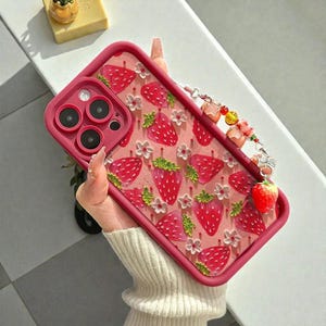 Puede incluir: Funda de teléfono rosa con un diseño de fresas y flores blancas. La funda tiene un borde rosa y un charm decorativo con cuentas y una fresa. Diseñada para proteger el teléfono. Medidas en centímetros.