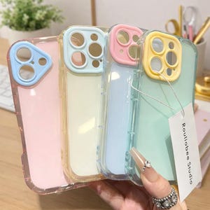 Puede incluir: Cuatro fundas de teléfono de colores pastel rosa, amarillo, azul y verde. Cada funda tiene una parte trasera transparente y un marco de color alrededor de la cámara y los bordes. Una etiqueta con el texto "Rouliabee Studio" está adjunta.