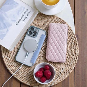May include: A light blue and clear phone case with a white magnetic charger and a pink quilted phone case are displayed on a woven mat. A small bowl of raspberries and a book with the text "Learn to Fly grow your wings" are also present.