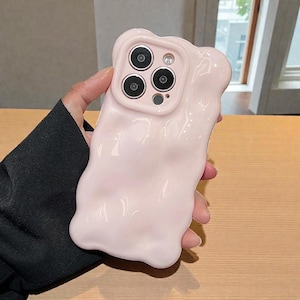 May include: A light pink phone case with a unique, wavy design. The case has a glossy finish and features a camera cutout with four circular lenses. The case is held by a person.