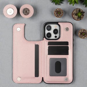 Puede incluir: Una funda de teléfono de cuero rosa claro con un borde negro, abierta para revelar ranuras para tarjetas y una ventana de identificación transparente. Dos soportes magnéticos rosas a juego son visibles arriba. La funda está diseñada para guardar un teléfono y tarjetas.