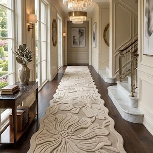 May include: A long, beige runner rug featuring a raised, carved floral pattern with large, flowing petals. The rug has an irregular, wavy edge and is positioned centrally along a dark wood floor in a hallway with white walls and gold lighting.