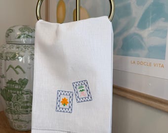 May include: White linen hand towel with embroidered floral and geometric designs, hanging on a gold-toned metal towel rack. The towel features a decorative hem and is displayed in a well-lit setting.