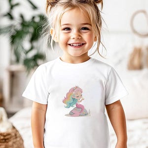 May include: A white t-shirt featuring a colorful illustration of a mermaid sitting on a rock. The mermaid has rainbow hair and a pink tail. The shirt is worn by a young girl with blonde hair.