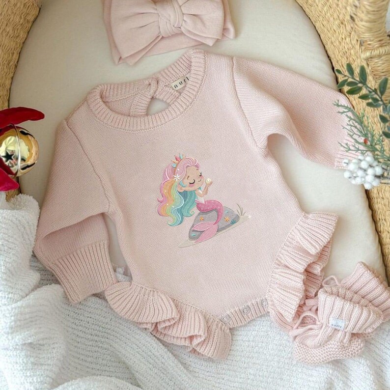 May include: A light pink baby outfit set, including a romper with a mermaid graphic, a matching headband, and booties. The romper features long sleeves and a ruffled hem. The mermaid has colorful hair and is sitting on a rock. The set is made of a soft knit material.