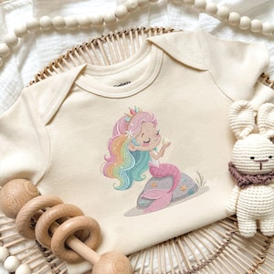 May include: Cream-colored baby bodysuit featuring a colorful mermaid illustration. The mermaid has rainbow hair, a pink tail, and a crown. She is holding a pearl. The bodysuit is made of soft fabric and is designed for infants.