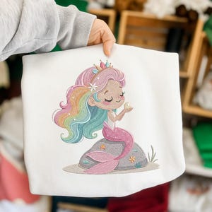 May include: White sweatshirt featuring an embroidered mermaid design. The mermaid has rainbow hair, a pink tail, and is holding a pearl. She is sitting on a gray rock with starfish and seaweed accents. The design is colorful and whimsical.