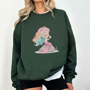 May include: Dark green sweatshirt featuring a cartoon mermaid with rainbow hair, a pink tail, and a pearl. The mermaid is sitting on a rock with starfish. The sweatshirt has a white undershirt.
