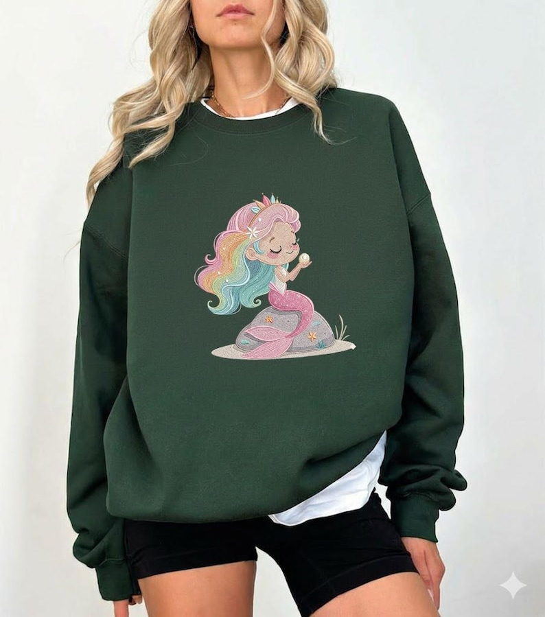 May include: Dark green sweatshirt featuring a cartoon mermaid with rainbow hair, a pink tail, and a pearl. The mermaid is sitting on a rock with starfish. The sweatshirt has a white undershirt.