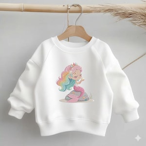 May include: White sweatshirt featuring a colorful mermaid illustration. The mermaid has rainbow hair, a pink tail, and a crown. The graphic is on a white, long-sleeved sweatshirt, perfect for casual wear.